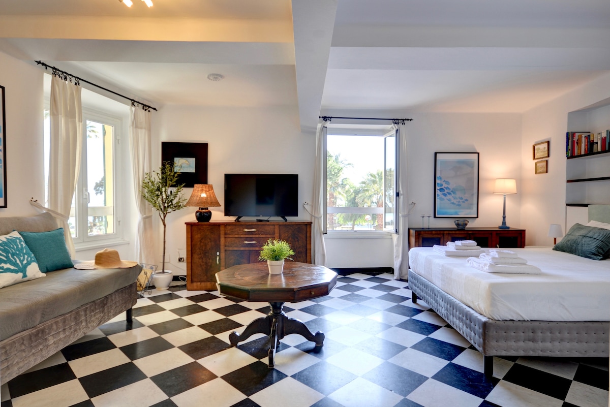 An airy bedroom features a spacious bed with fresh linens alongside a single sofa bed. A flat-screen TV is mounted on the wall, and natural light floods in through large windows. The room is decorated with elegant furnishings and a contrasting black-and-white tiled floor.