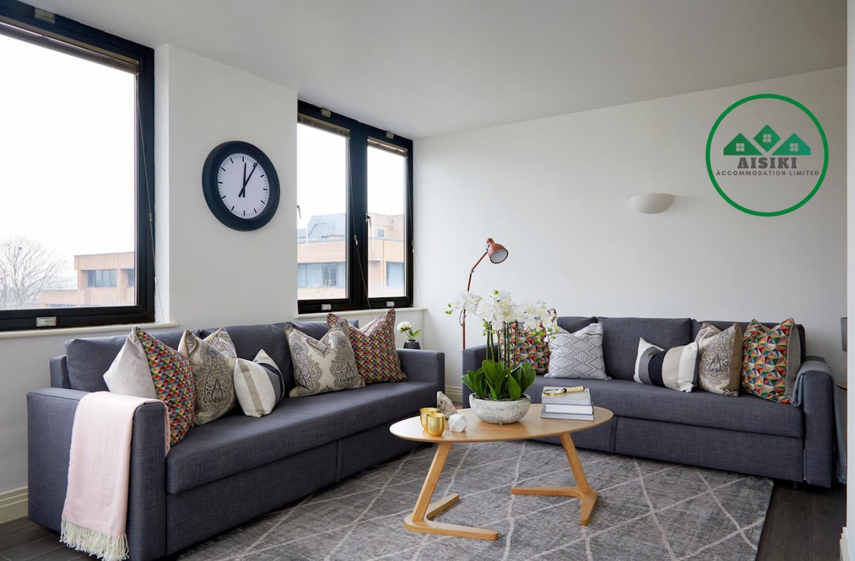 A cozy living area features two comfortable sofas adorned with decorative cushions. A round coffee table sits centrally on a patterned rug. Large windows allow natural light to brighten the space, enhancing the simple and modern decor.