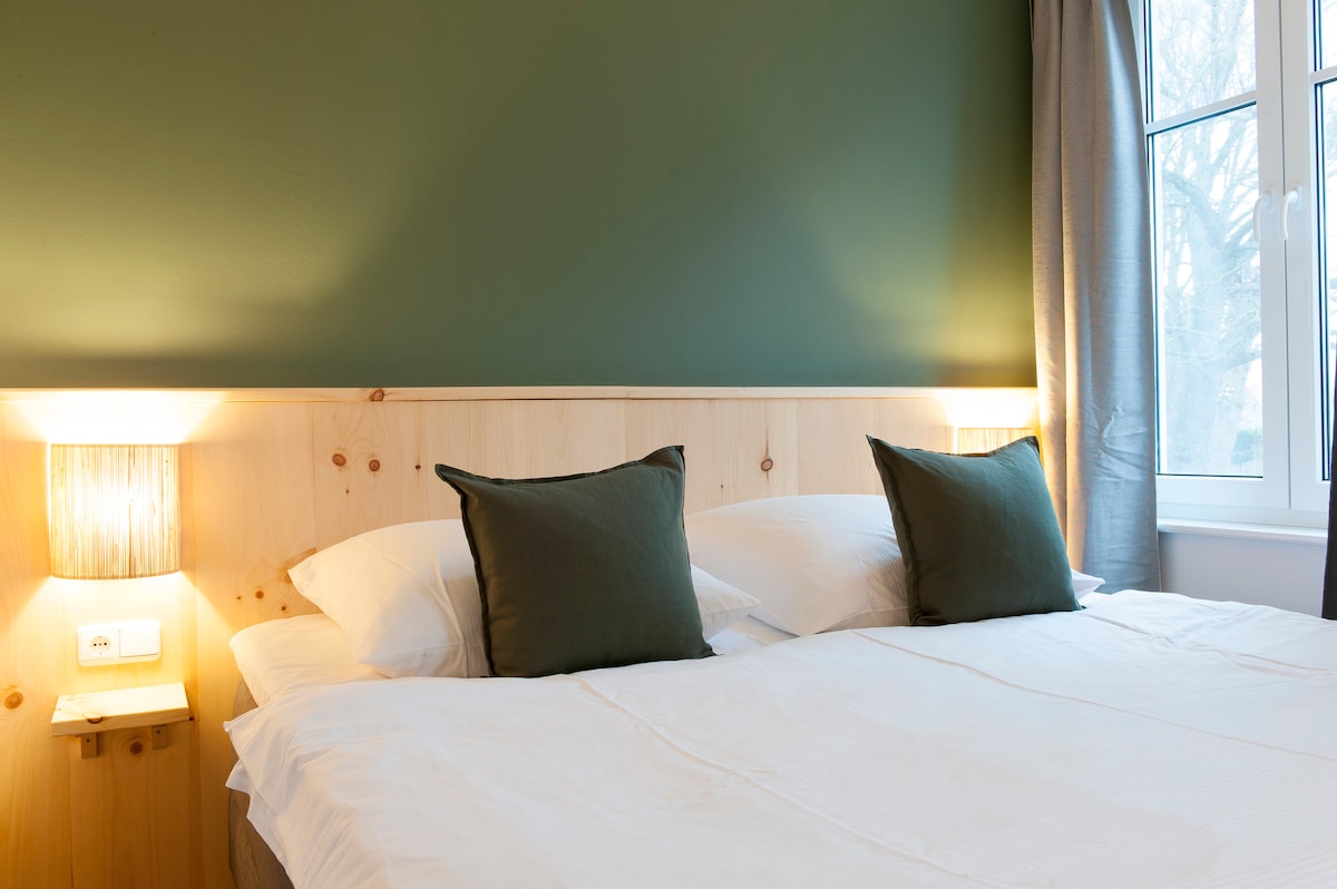A cozy sleeping area features a spacious bed with white linens and green accent pillows. The light wood headboard enhances the calming atmosphere, complemented by bedside lamps. Natural light enters through a nearby window, draped with light grey curtains.