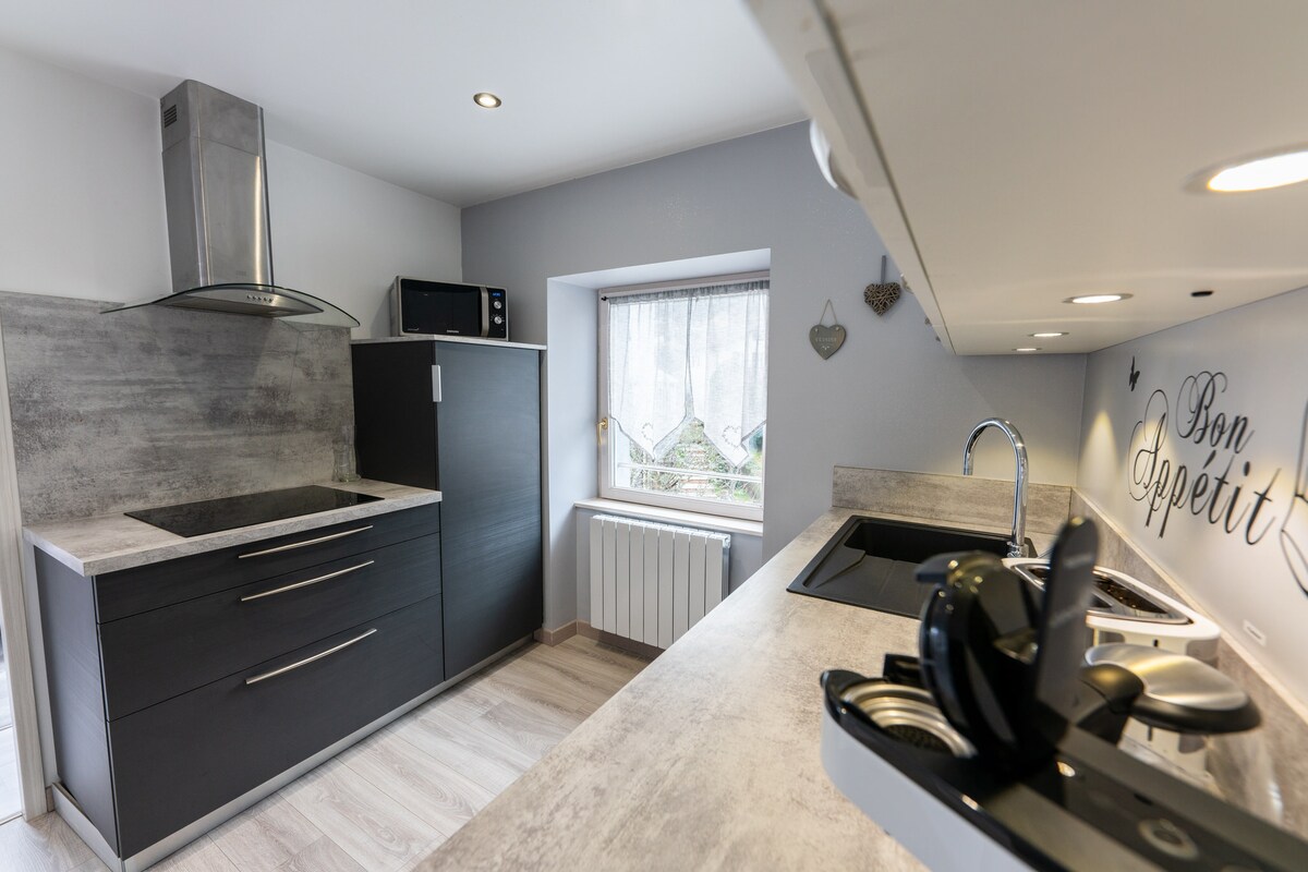 A modern kitchen is featured with sleek dark cabinetry and a light-colored countertop. Essential appliances such as a microwave and a stovetop are prominently displayed. A window allows natural light to illuminate the space, along with decorative text on the wall.