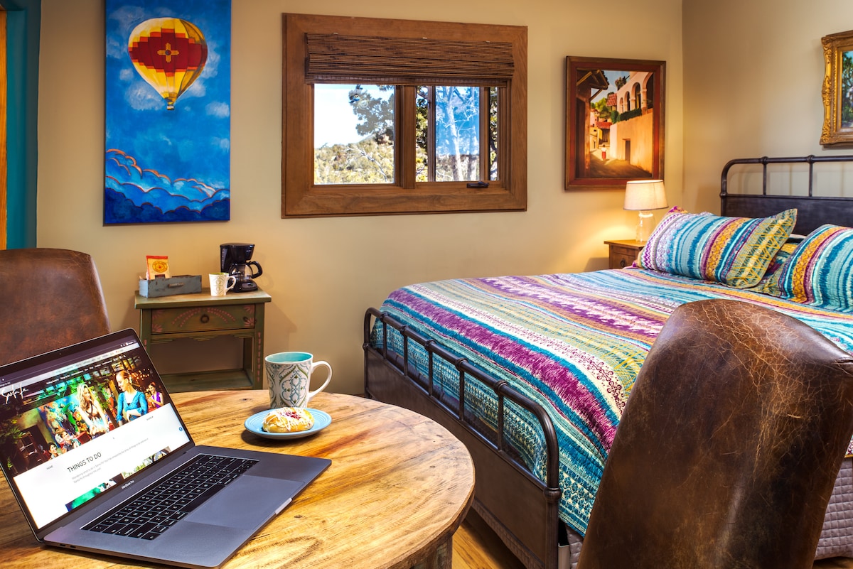 A vibrant room showcases a king-sized bed adorned with a colorful bedspread. A wooden table and chairs offer a cozy dining space. Natural light filters through a window with views of the surrounding landscape. A laptop and coffee cup are placed on the table, indicating a comfortable working area.