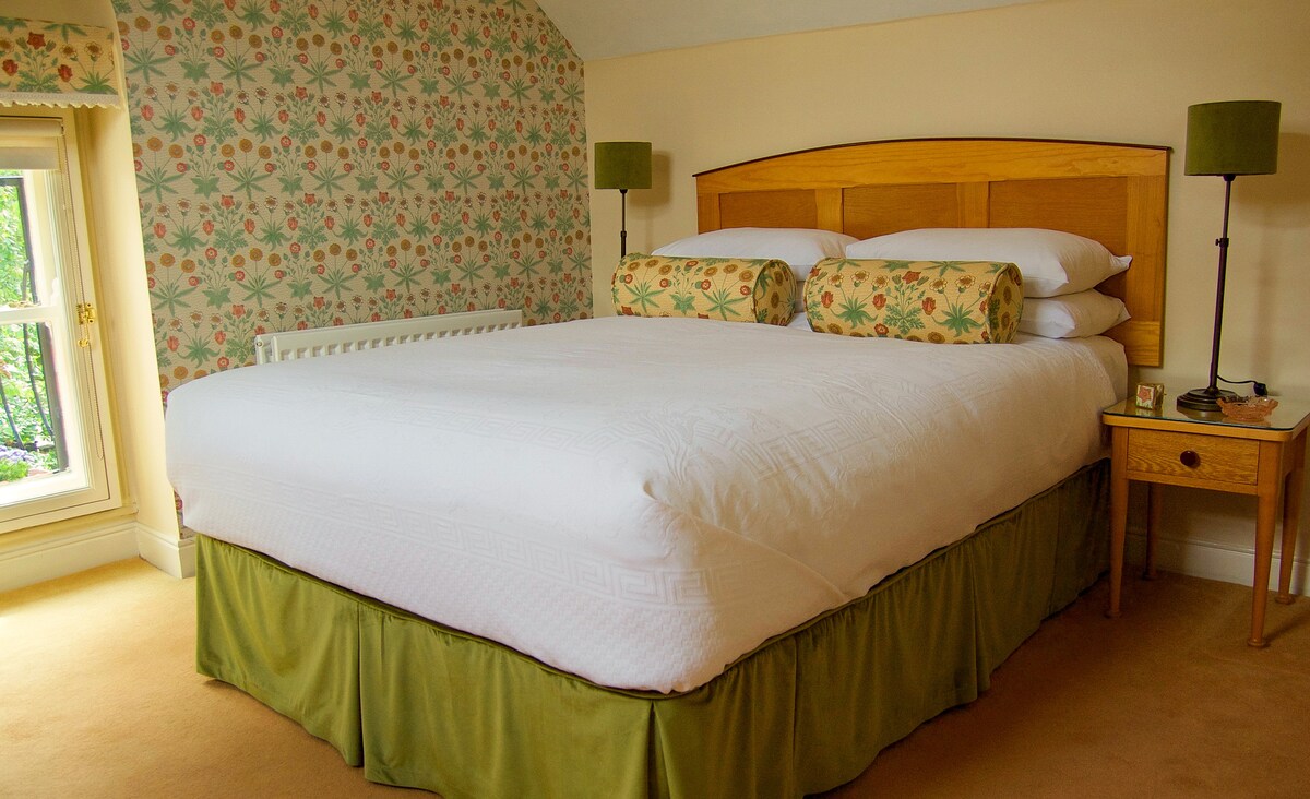 A spacious bedroom features a large upholstered bed with decorative cushions and soft bedding. Floral wallpaper adds a touch of color, while two bedside lamps provide gentle illumination. A window allows natural light to enter, enhancing the inviting ambiance of the room.