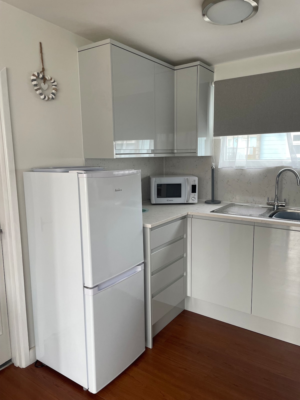 A bright kitchen area features sleek white cabinetry and a compact design. A refrigerator, microwave, and stainless-steel sink are prominently displayed. The countertops are light-colored, and natural light filters in through a window covered by a roller shade.