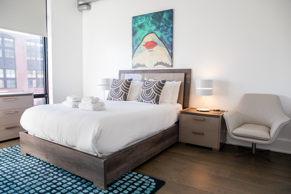 A spacious bedroom features a comfortable bed adorned with crisp white linens and decorative pillows. Two bedside lamps provide soft lighting beside the bed. A stylish piece of artwork hangs above, complemented by a textured area rug and a sleek armchair, creating a serene atmosphere.