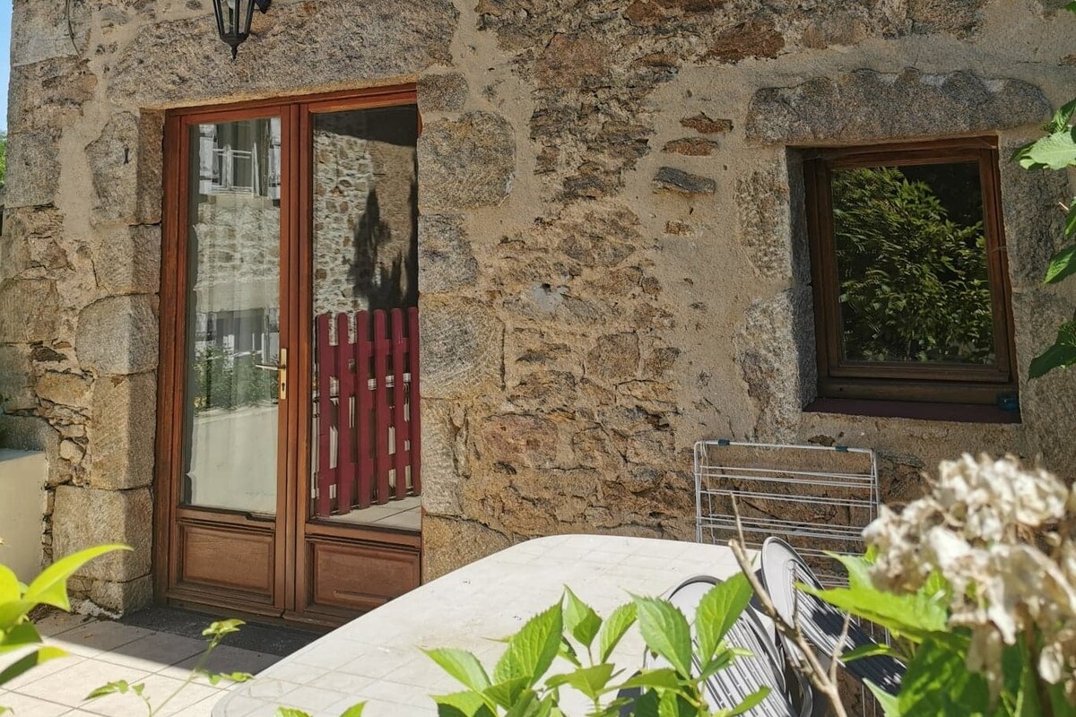 An outdoor dining area features a table and chairs set against a rustic stone wall. Large glass doors reflect the surrounding greenery and allow natural light into the space. A small window provides additional views of the courtyard.