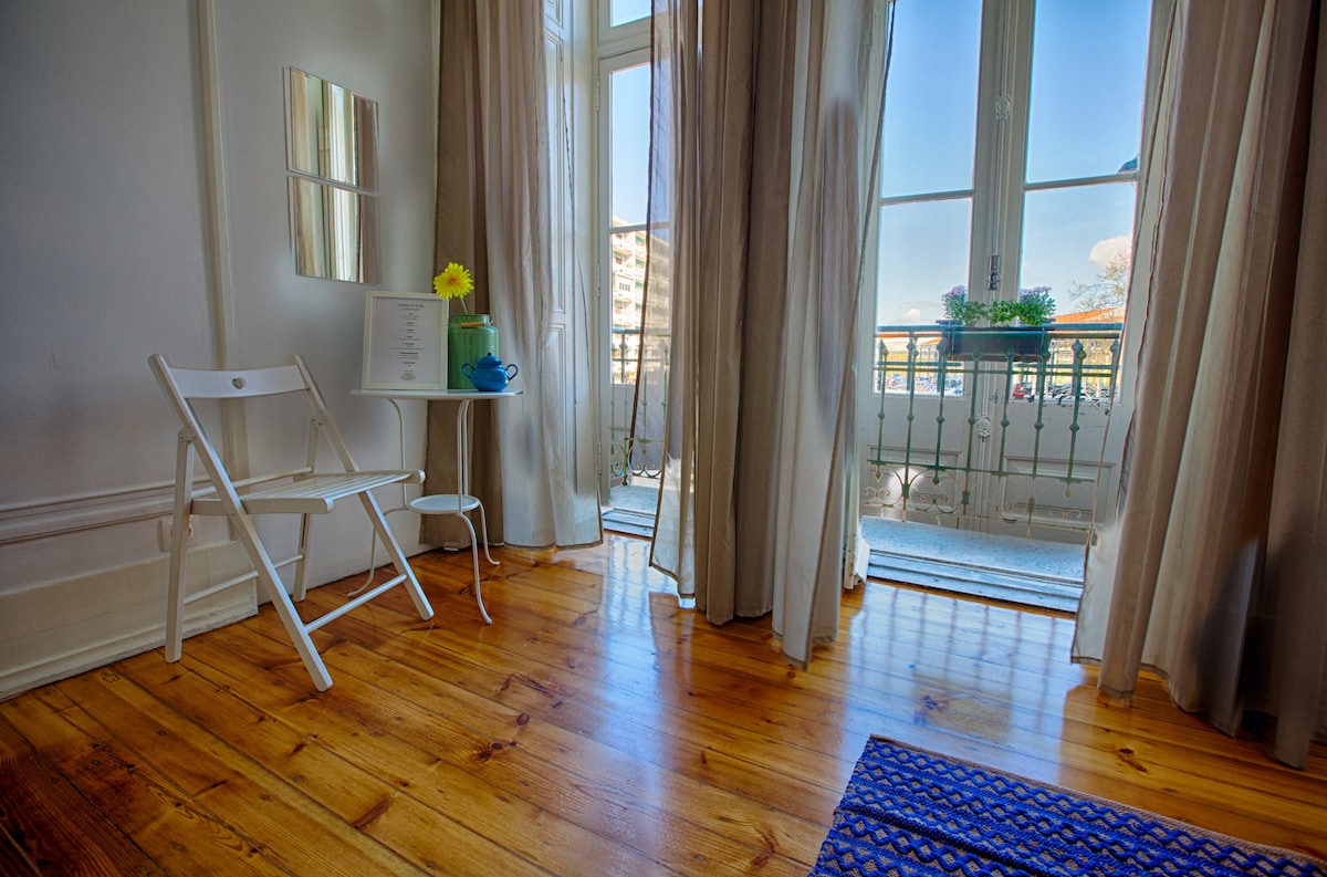 A bright room features light wooden flooring and large windows draped with sheer curtains. A small white table and folding chair are positioned near the window, adorned with a blue cup and a yellow flower. Natural light fills the space, enhancing its airy feel.