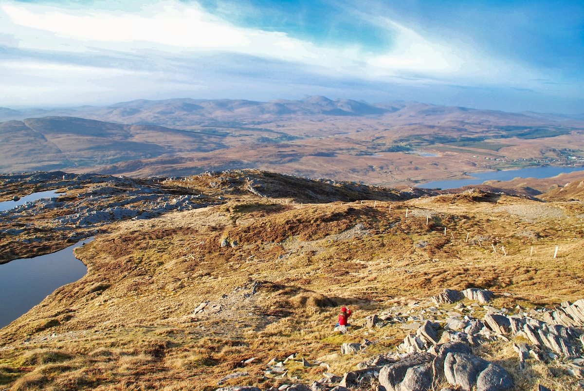 A panoramic view of rolling hills extends under a clear blue sky, featuring a mix of grassy terrain and rocky outcrops. Small bodies of water are visible in the landscape, adding natural beauty to the serene environment. A lone figure is walking along a path, enhancing the sense of scale.