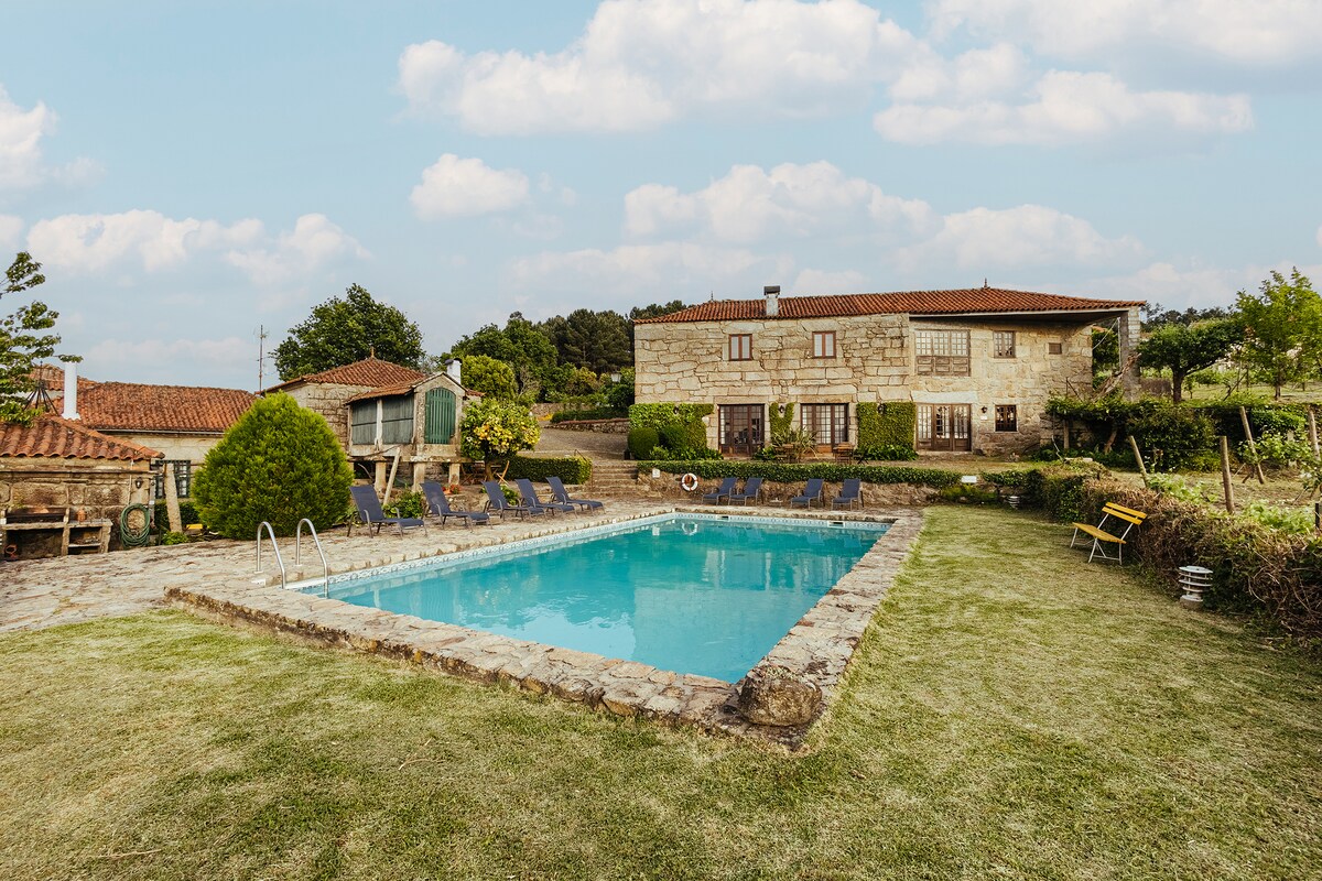 An outdoor swimming pool is surrounded by a stone patio and neatly arranged lounge chairs. Beyond the pool, a rustic stone building features windows and a red-tiled roof, set amidst greenery and landscaped gardens.