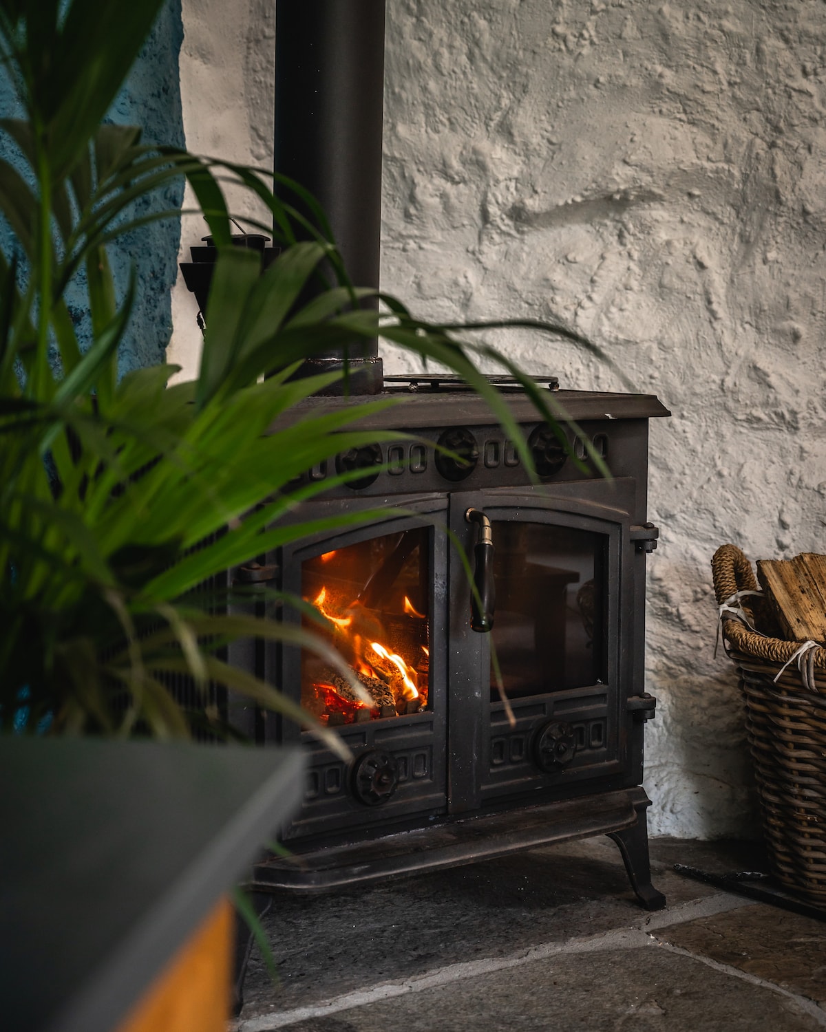 A traditional log burning stove is positioned against a textured stone wall, with flames visible through its double doors. Nearby, a woven basket is filled with firewood, and a green plant adds a touch of nature to the warm setting.