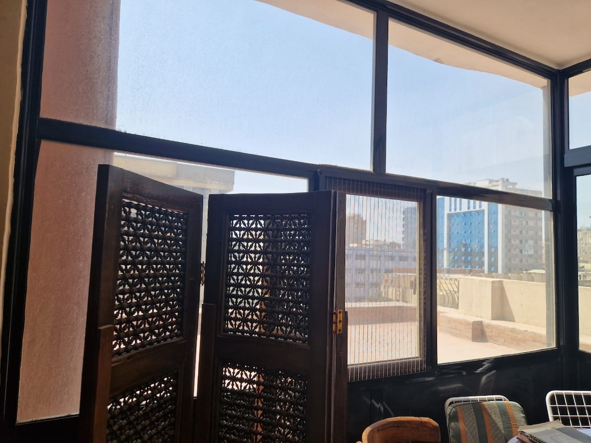 A sun-filled seating area is showcased with large windows that provide a panoramic view. Intricately designed wooden screens add a decorative element, while the soft light highlights the spaciousness of the room. Nearby chairs create a relaxed atmosphere suitable for enjoying the view.
