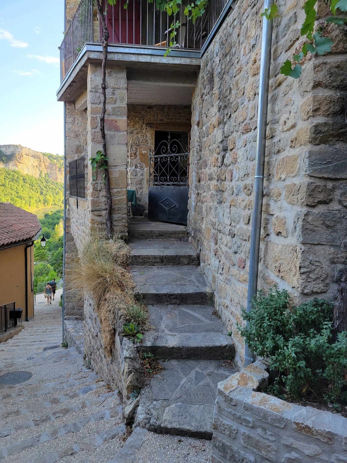 A narrow stone pathway leads to a black gate framed by an intricately designed metal grille. The pathway features uneven stone steps, bordered by greenery and rustic stone walls. Soft sunlight casts gentle shadows, highlighting the natural textures of the surroundings.