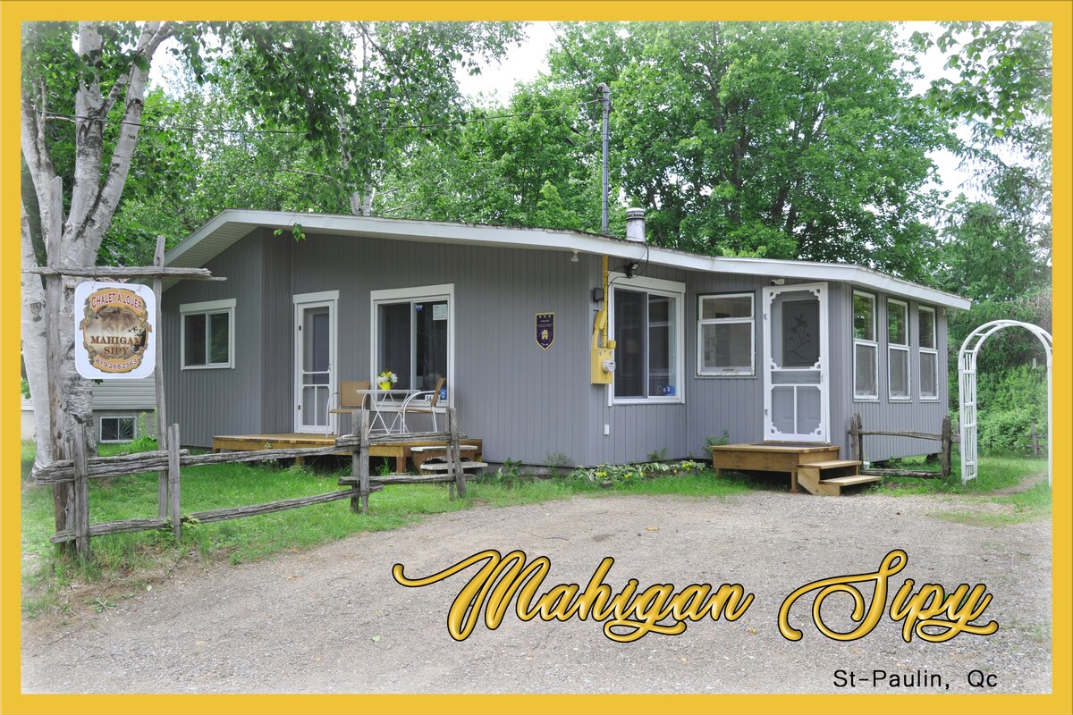 A gray chalet is situated in a serene natural setting, featuring a welcoming porch with outdoor seating. Large windows allow for ample light, and a sign reads ‘Mahingan Siq’ at the entrance, enhancing the charm of this peaceful retreat.