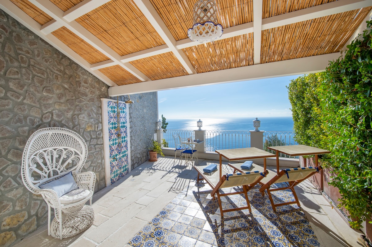 A spacious terrace is presented with a stunning sea view, featuring natural stone walls and a woven canopy. Comfortable seating includes a decorative chair and sun loungers. Patterned tiles add color to the floor, creating a serene outdoor space for relaxation.