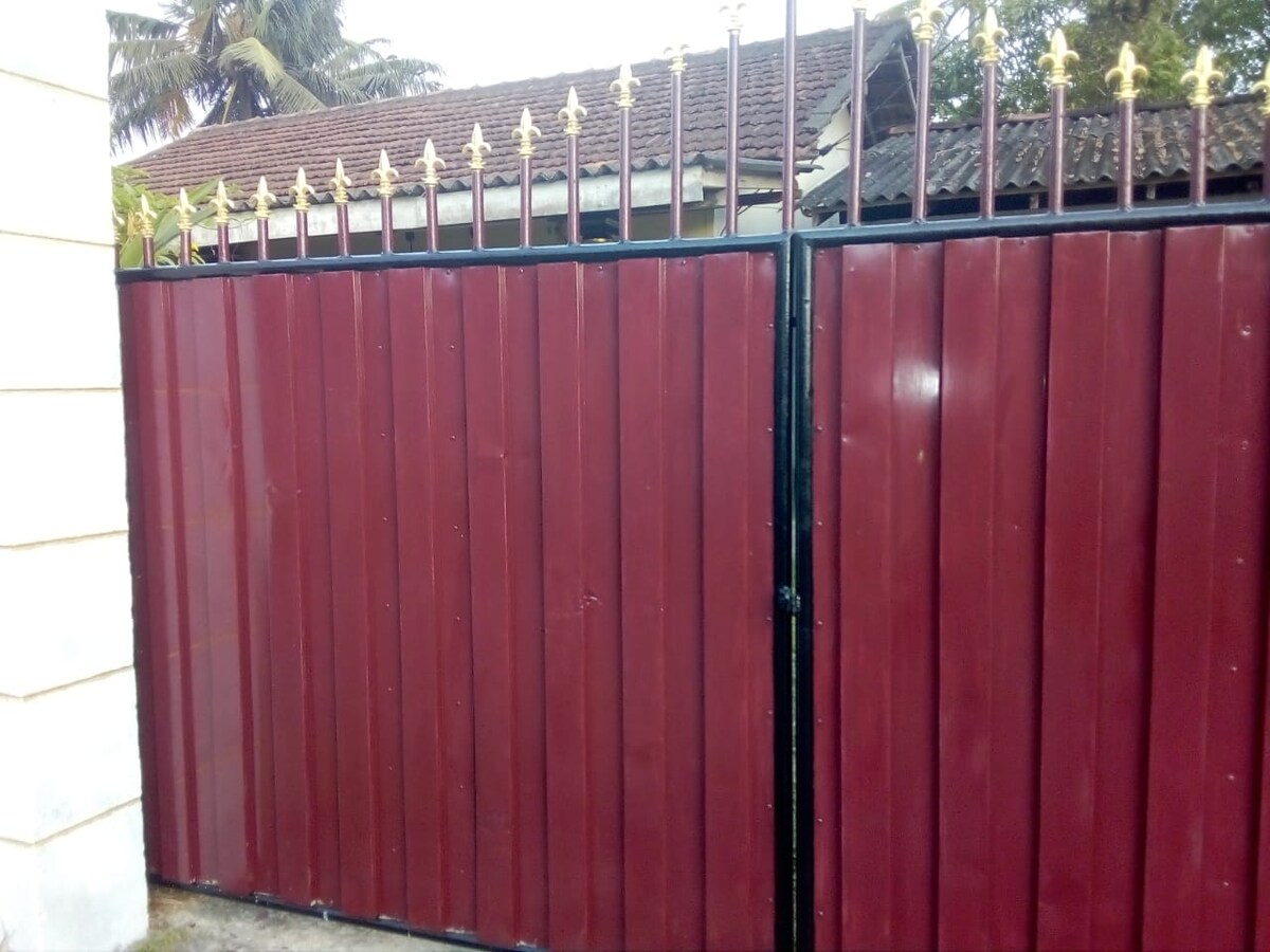 A gated entrance features a maroon vertical slat design with a decorative gold-tipped railing at the top. The gate is set against a backdrop of lush greenery, with additional structures partially visible behind it.