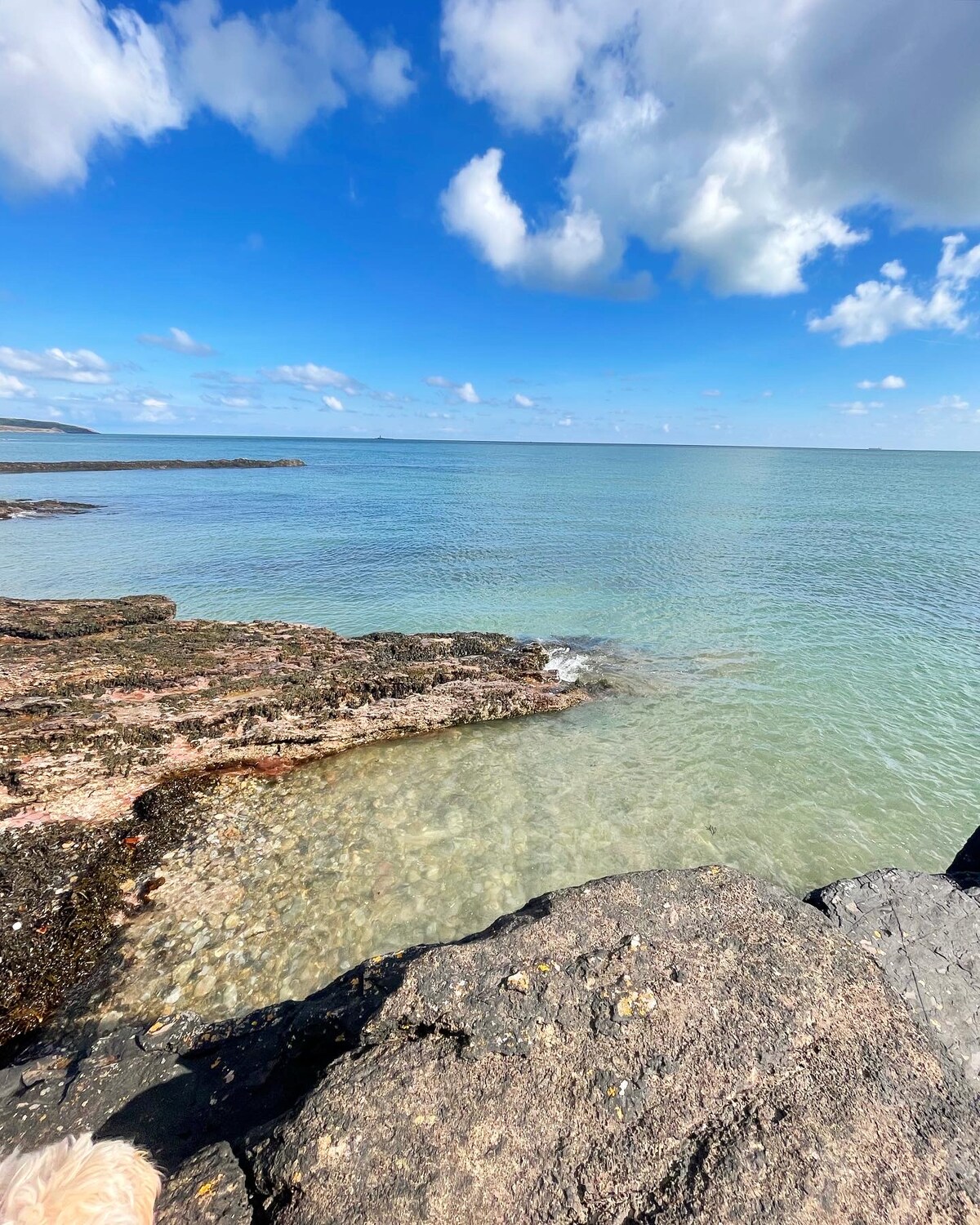 A tranquil coastal scene is depicted, showcasing clear waters gently lapping against rugged rock formations. The sky is bright with scattered clouds, and distant shorelines can be seen on the horizon, creating a serene and inviting atmosphere.