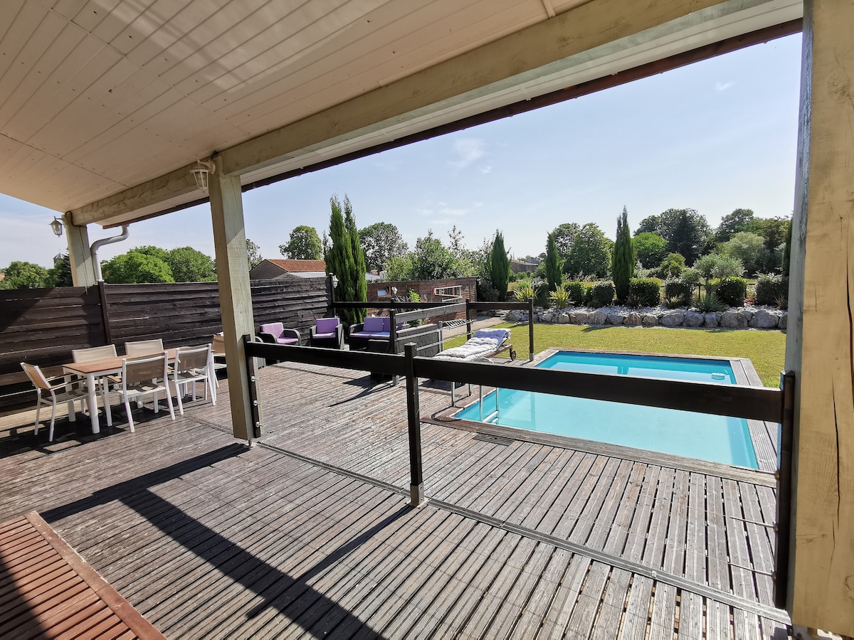 An expansive deck features a swimming pool surrounded by landscaped greenery. Comfortable seating areas with cushions are visible, along with a dining table set for outdoor meals. The modern villa’s structure provides a blend of shaded and sunlit areas, enhancing the outdoor living experience.