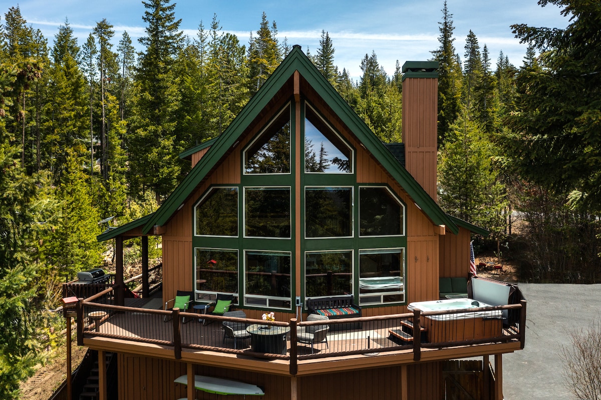 The chalet exterior features large windows, offering expansive views of the surrounding forest. A spacious deck is visible, equipped with lounge chairs and a hot tub, all set against a backdrop of towering trees under a clear blue sky.