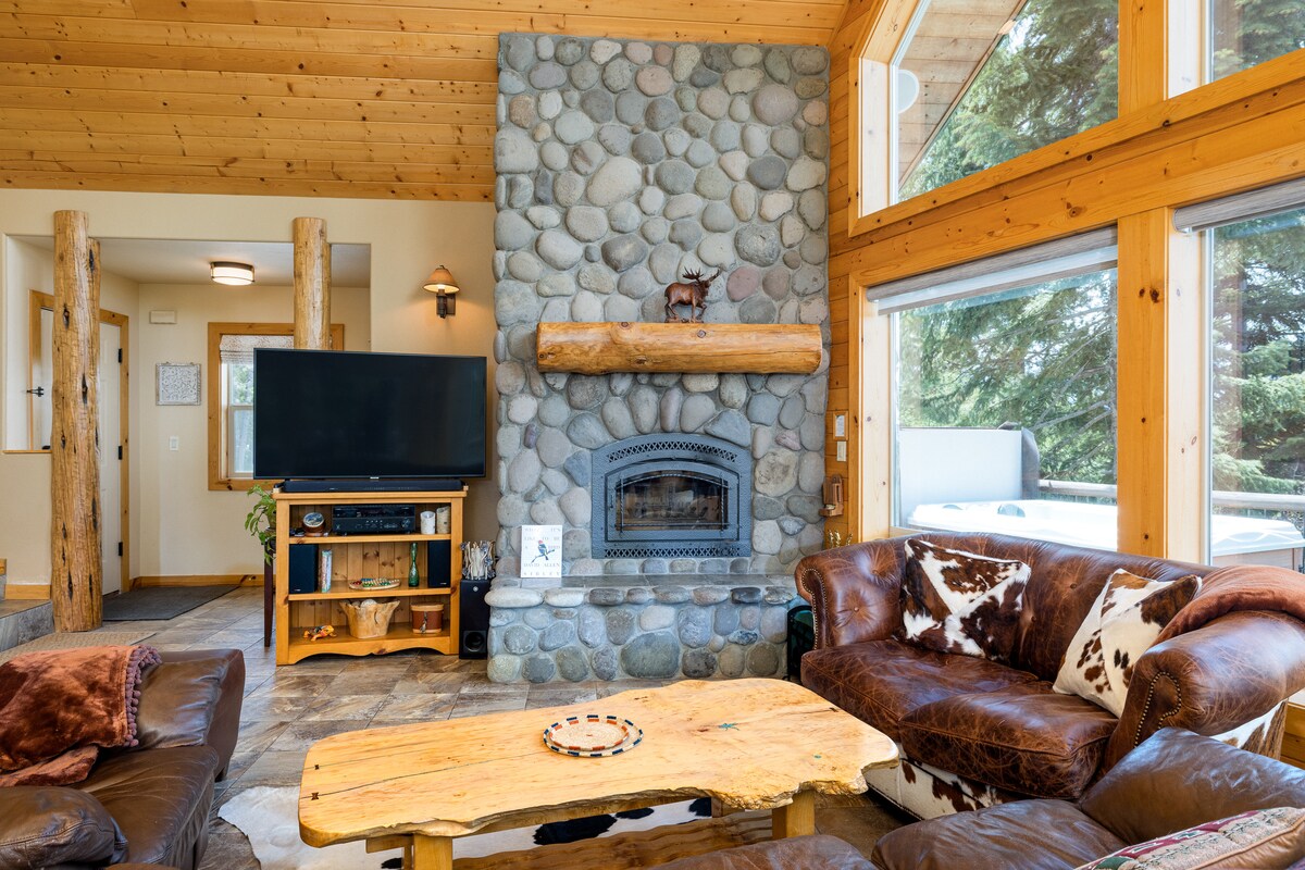 The living room features a large stone fireplace framed by natural wood beams. Comfortable leather sofas surround a rustic wooden coffee table, enhancing the cozy atmosphere. A flat-screen TV is positioned nearby, and expansive windows provide views of the surrounding wooded area.