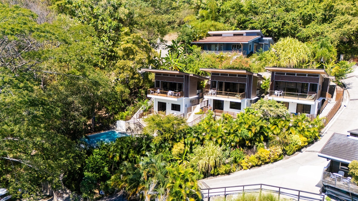 The image features a modern apartment complex nestled among lush greenery, with three units visible from an elevated perspective. Each unit includes a spacious balcony, and a shared infinity pool is set within the tropical landscape, complementing the natural surroundings.