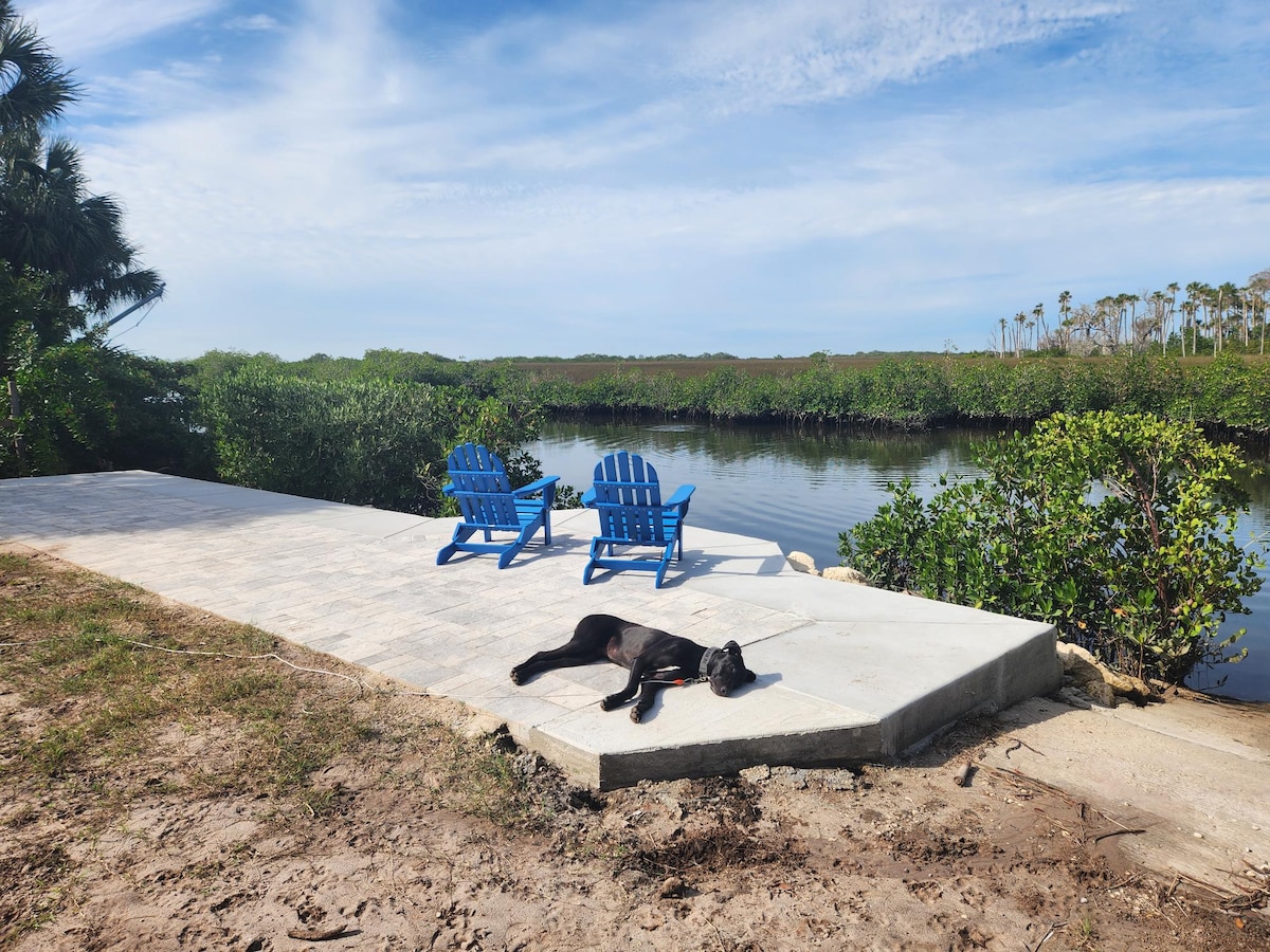 Two blue adirondack chairs are positioned on a stone patio, overlooking a calm canal lined with lush vegetation. A black dog is lying comfortably on the sunlit patio, enjoying the serene surroundings under a clear blue sky.
