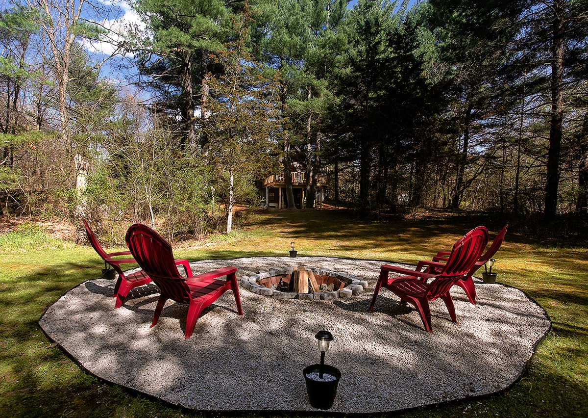 An outdoor fire pit area features four red lounge chairs arranged around a circular gravel space. Soft lighting from nearby fixtures enhances the peaceful surroundings, while tall trees provide a natural backdrop, creating a secluded atmosphere for relaxation.