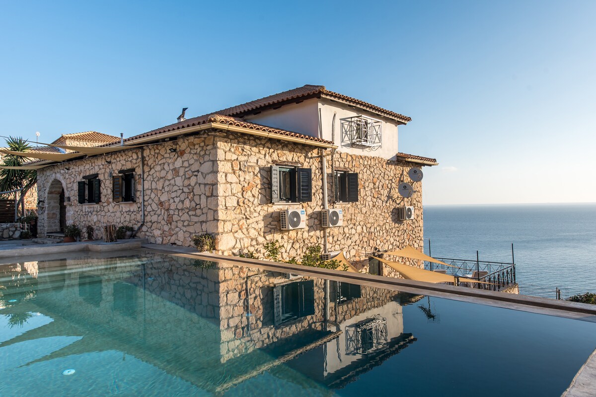 A stone building is positioned near the water's edge, featuring large windows that reflect the blue sky. An infinity pool extends towards the horizon, providing a seamless transition to the sea. Air conditioning units are visible on the exterior, indicating a comfortable atmosphere.