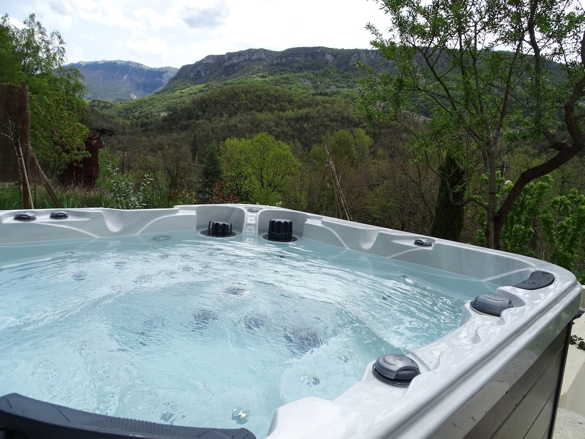 An outdoor spa is situated with a view of rolling hills and greenery. The bubbly water glistens under the natural light. Lush trees and distant mountains form a serene backdrop, providing a peaceful setting for relaxation.