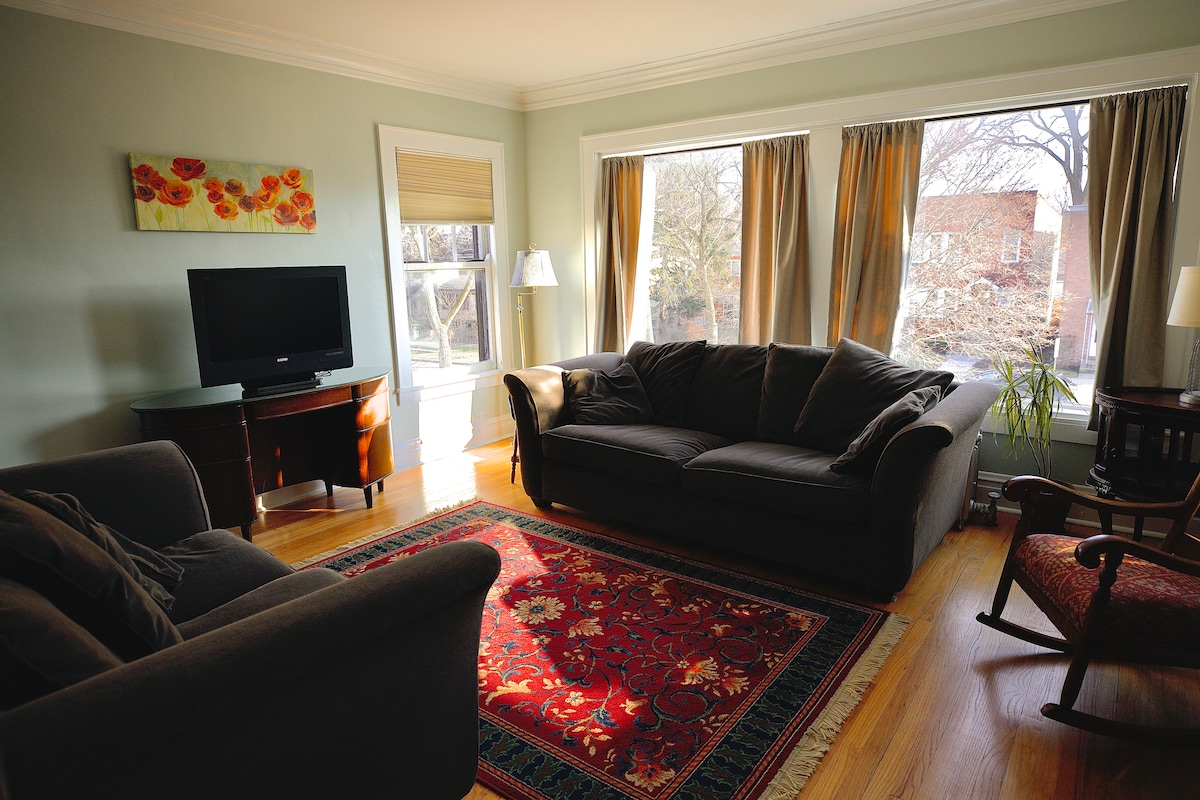 A spacious living room features large windows draped with light curtains, allowing natural light to fill the space. A comfortable sofa and loveseat are positioned around a decorative area rug, with a television mounted on a cabinet. A rocking chair adds additional seating.