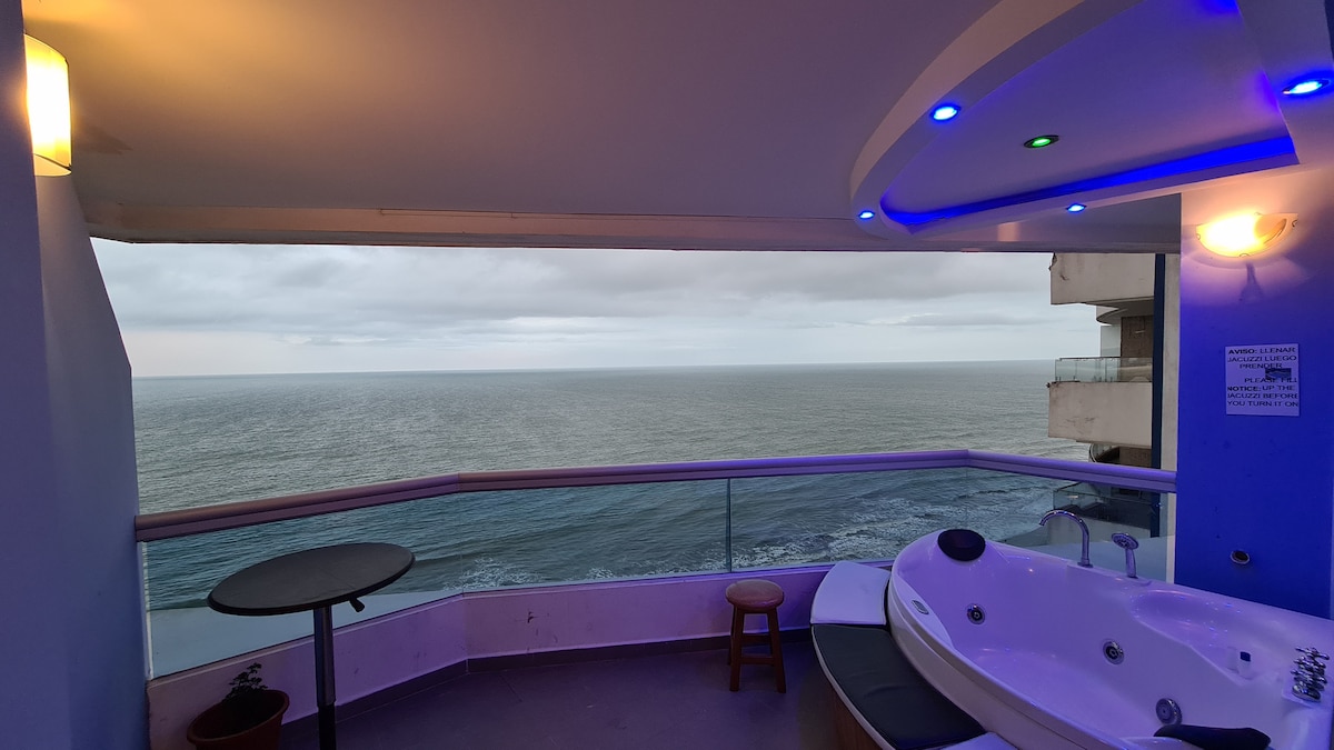 The balcony presents a private jacuzzi and a small table accompanied by a stool, offering views of the ocean horizon. Soft LED lights create a gentle ambience along the edge, while the glass railing provides unobstructed sightlines to the sea.