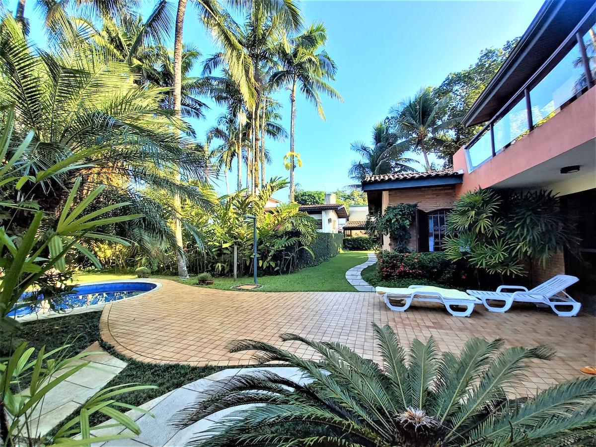 A tropical garden is showcased, featuring lush palm trees surrounding a private swimming pool. A paved area provides space for outdoor seating, while a few sun loungers are positioned nearby. The house is partially visible, blending harmoniously with the natural landscape.