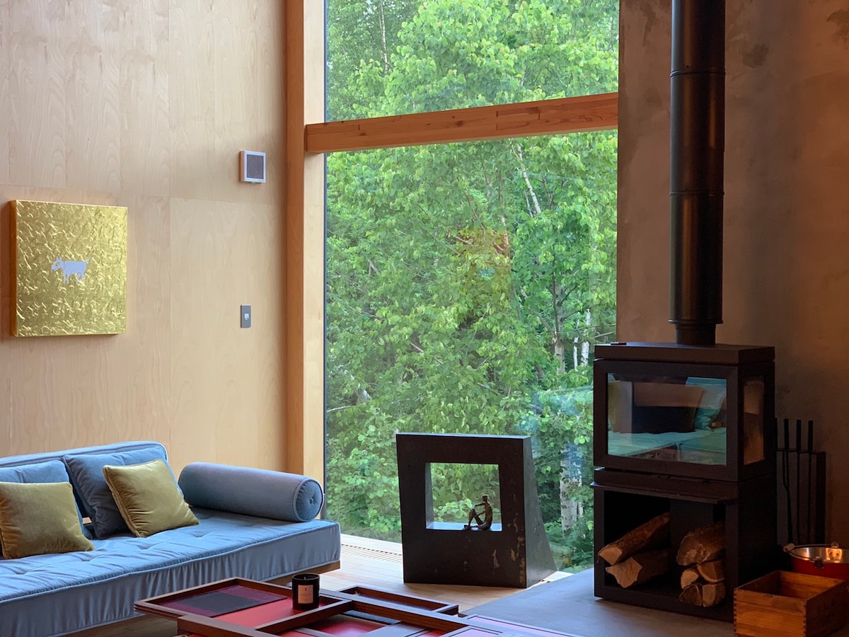 A modern living area features a blue sofa with decorative cushions, positioned near large windows that frame a view of lush greenery. An elegantly designed wood stove stands beside a stack of firewood, enhancing the cozy ambiance of the space.