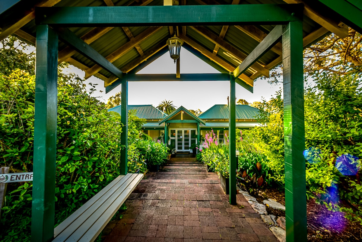 An inviting entrance features a covered walkway adorned with greenery, leading to large glass doors that open into the guesthouse. A bench sits alongside the path, creating a welcoming space surrounded by vibrant plants and flowers.