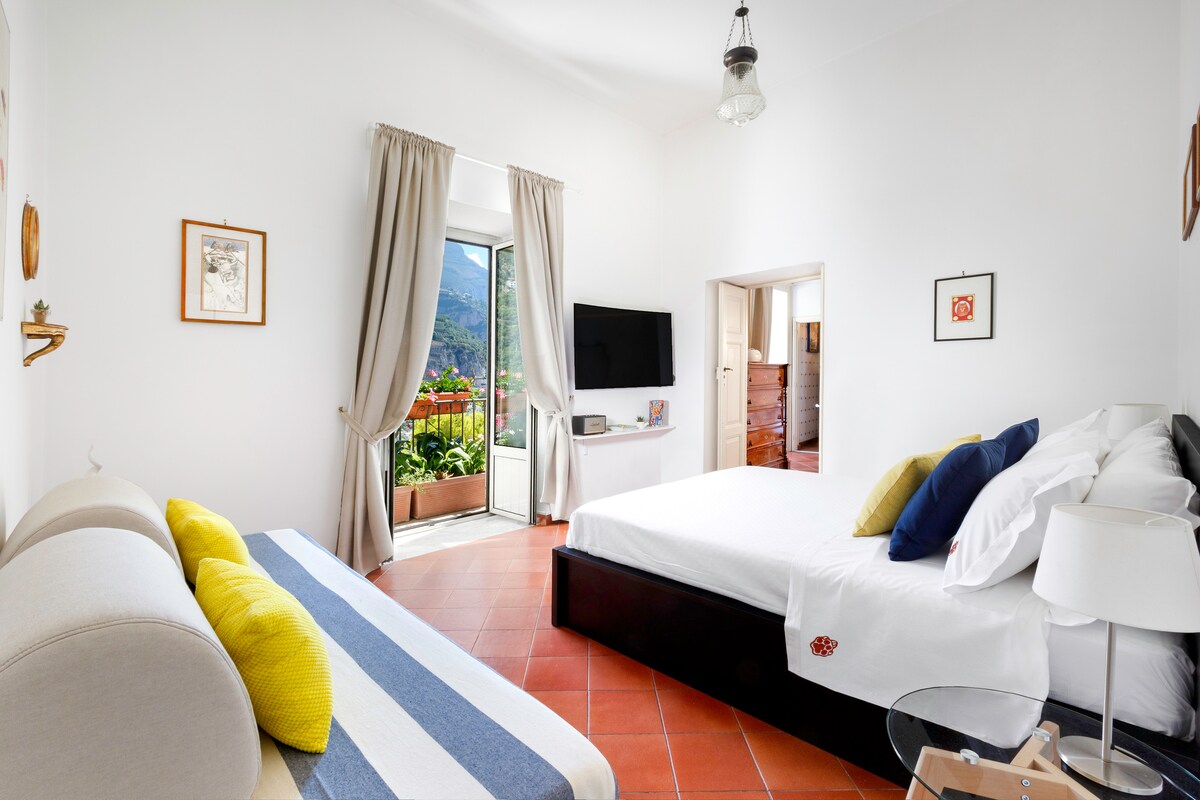 A bright and spacious bedroom features a king-sized bed with white linens and decorative pillows. A sleeper sofa with blue and yellow accents sits beside a glass door that opens to a balcony. A large television is mounted on the wall, offering modern amenities.