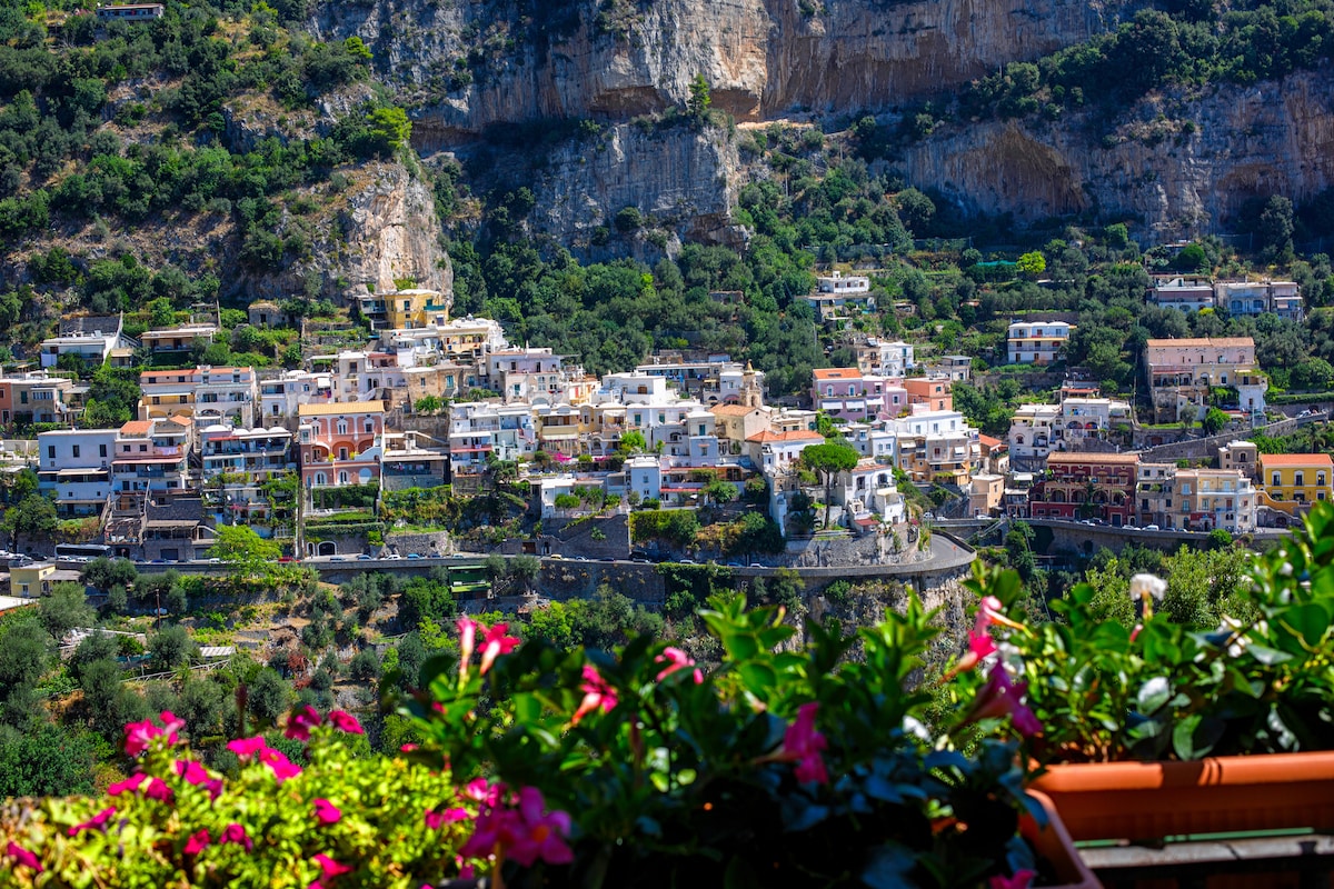 A vibrant view of Positano is captured, showcasing colorful buildings nestled into the hillsides, framed by lush greenery. The rocky cliffs rise majestically in the background, complementing the charming architecture of the town. Flowering plants provide a lively foreground to the scene.