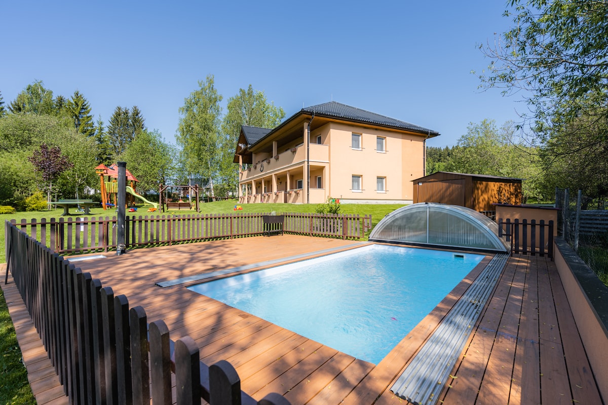 An inviting outdoor area features a rectangular pool, surrounded by a wooden deck and fenced for privacy. A two-story building stands in the background, complemented by lush greenery and children's play equipment. The space offers a sense of openness and tranquility.