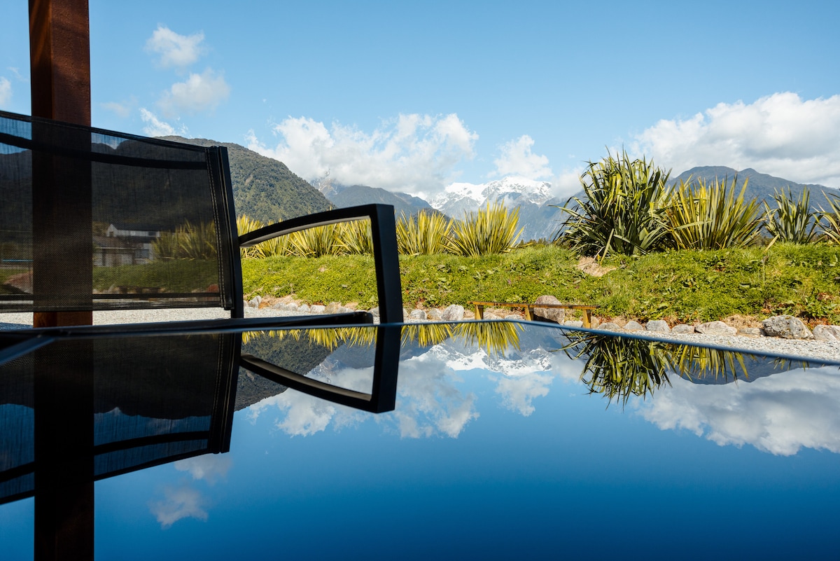 A tranquil scene is captured with a still surface reflecting the surrounding mountains and sky. Lush greenery frames the area, contrasting with the serene blue of the water, while a chair's silhouette hints at relaxation and enjoyment of the view.