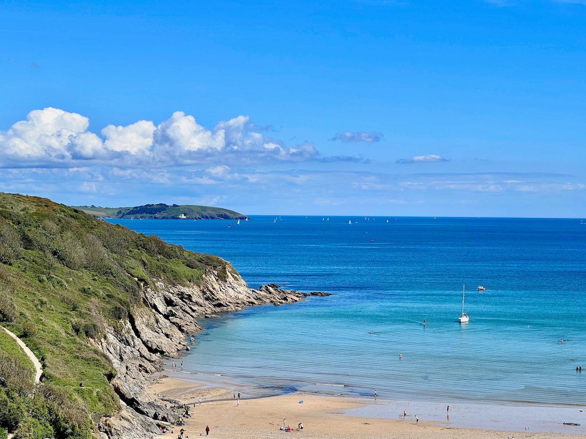 An expansive view of Maenporth Beach reveals soft golden sands meeting clear turquoise waters. Gentle waves lap at the shore, while a few small boats are seen in the distance against a backdrop of rolling hills and a bright blue sky dotted with fluffy clouds.