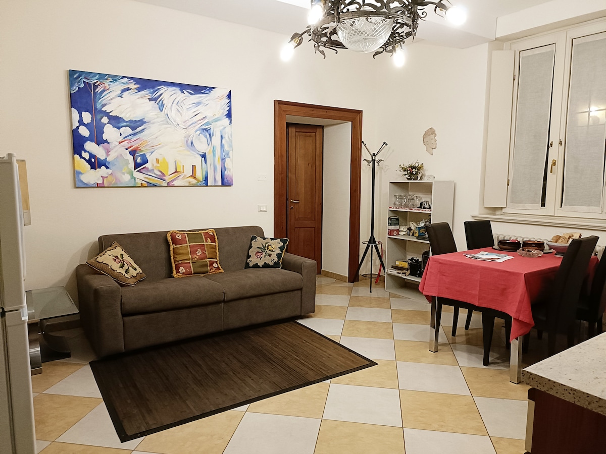The living area features a comfortable sofa in neutral tones with decorative cushions and a colorful artwork mounted on the wall. A dining table set for four sits nearby, complemented by dark chairs. Natural light enters through the window, which is covered by light-colored curtains.