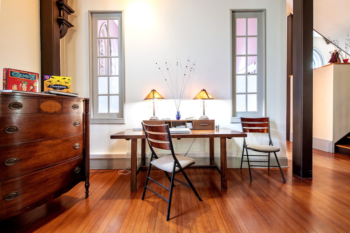 A workspace is set up in a historic setting, featuring a long wooden table with two chairs. Natural light filters through two tall windows, illuminating the room. A vintage dresser adds character, complemented by two decorative lamps on the table.