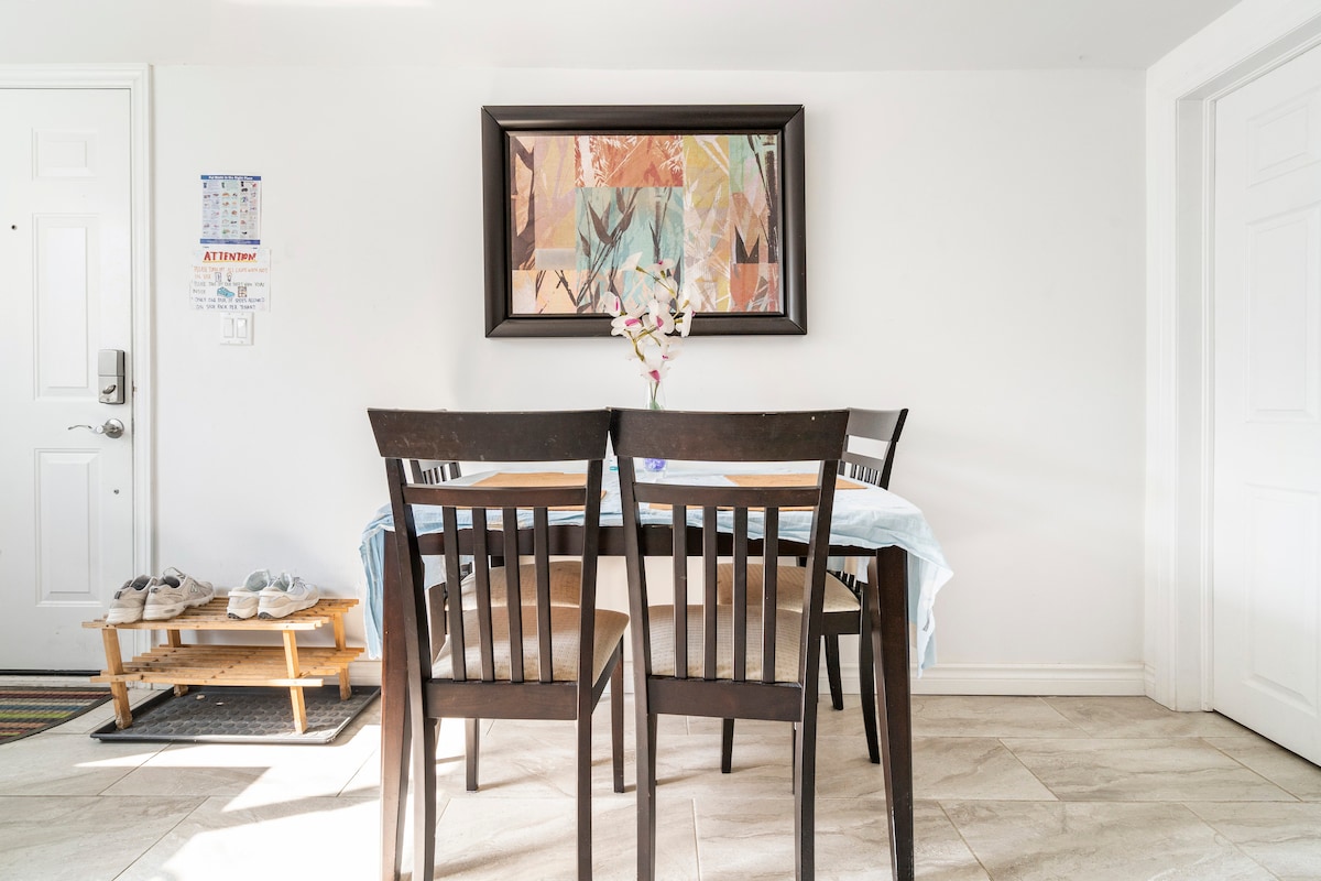 A dining area features a wooden table surrounded by four chairs, accented by a floral centerpiece. A colorful piece of wall art hangs above the table, while a small shoe rack and door are visible in the background, enhancing the room's functional layout.