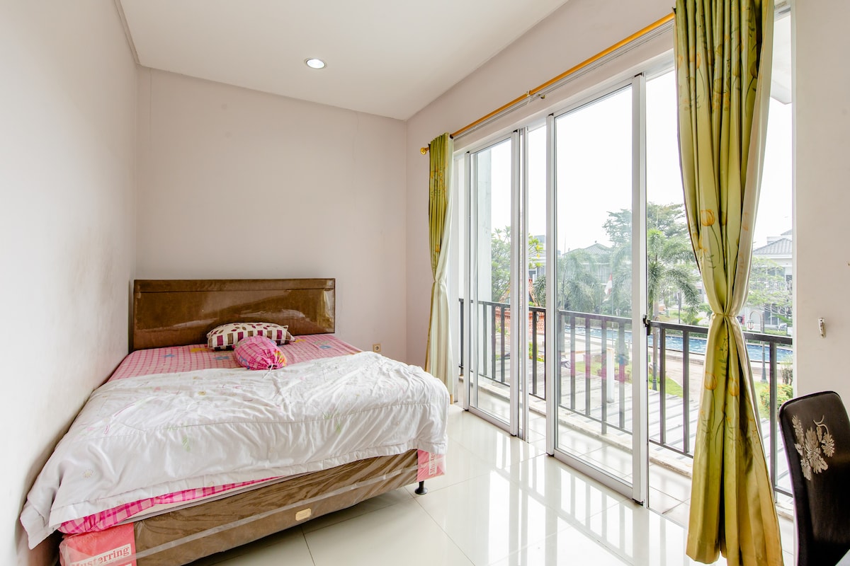 A private room features a spacious bed with striped bedding, accompanied by a pink accent pillow. Large glass doors provide an abundance of natural light and offer access to a balcony with green views. Bright green curtains frame the windows, adding a refreshing touch.