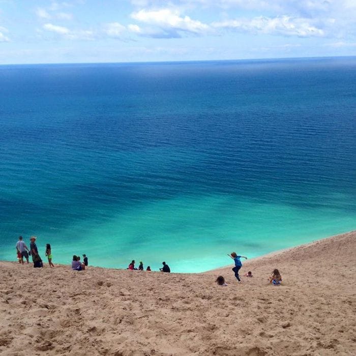 An expansive sandy dune overlooks a vibrant blue-green lake under a clear sky. Several individuals are seen enjoying the beach, with a few people climbing the dune while others sit on the sandy surface, taking in the serene views.
