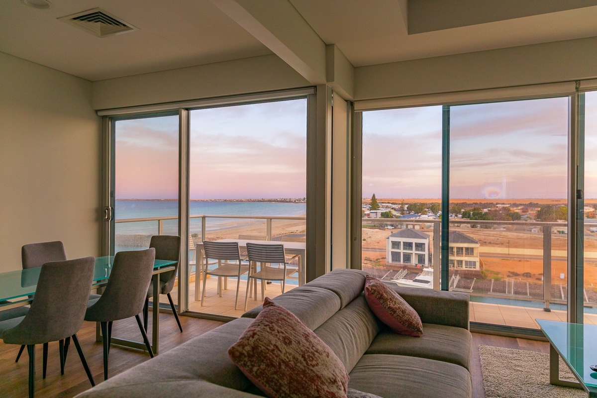 A modern living space features large glass doors that provide expansive views of the ocean and town. A comfortable sofa is positioned in the foreground, while a dining area is set with a glass table and several chairs. Soft lighting enhances the room's inviting ambiance.