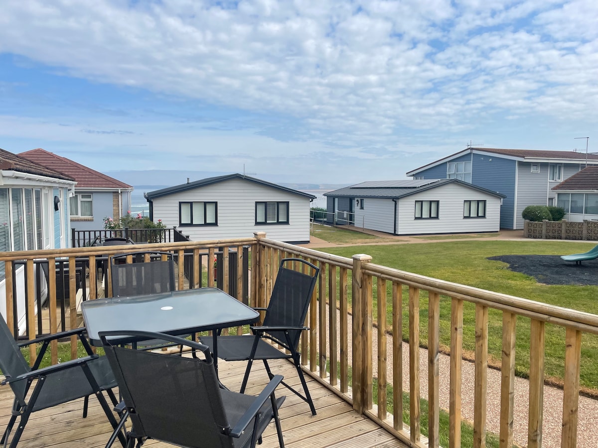 A wooden decked area is shown, featuring a table with four chairs designed for outdoor dining. Nearby, other cottages can be seen, with well-maintained lawn and distant ocean visible under a partly cloudy sky.