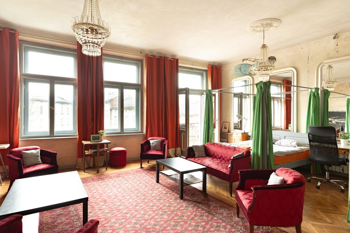 A spacious living area features large windows adorned with red drapes, allowing ample natural light. Elegant chandeliers hang from the ceiling, while a mix of plush seating, including red armchairs and a sofa, creates an inviting space. A desk and bed are visible in the background.