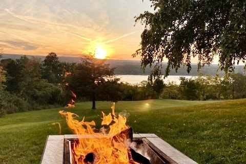 A fire pit is featured prominently in the foreground, with flames dancing above logs. In the background, a sunset casts warm light over a grassy landscape, and a body of water is visible, framed by trees and rolling hills.