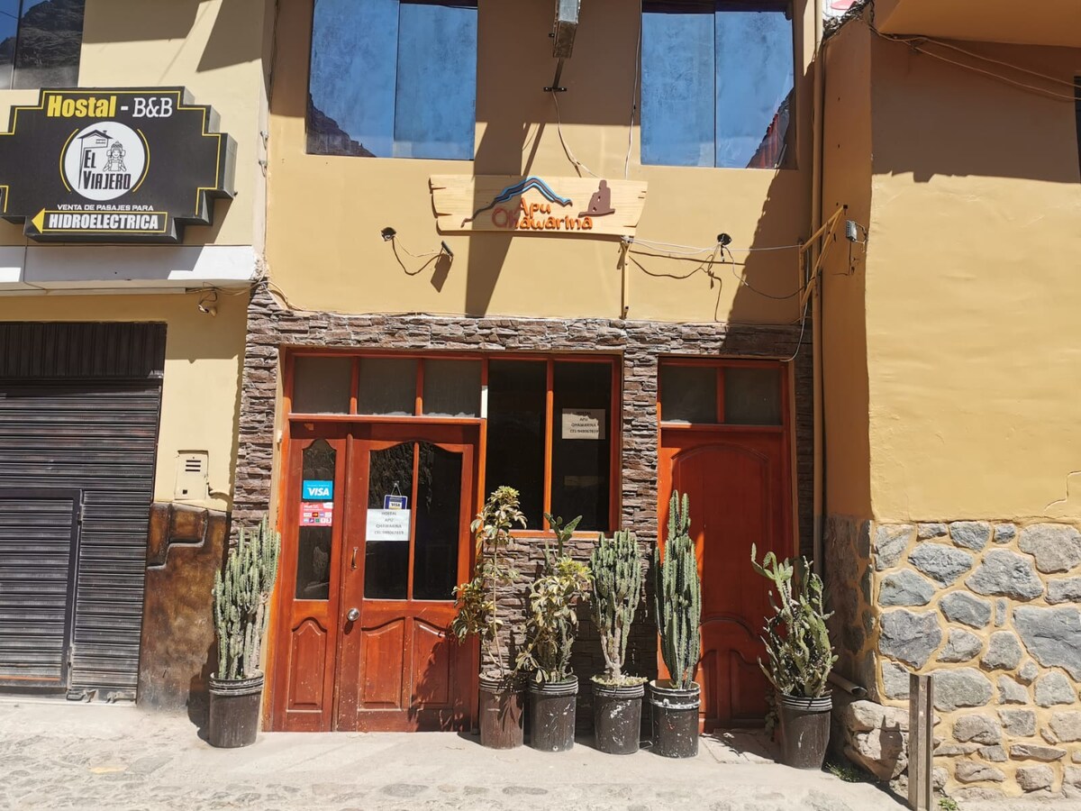 The hostel's entrance features wooden doors framed by stone accents. Planters with cacti are positioned on either side, adding a natural touch. A sign above the doors indicates the hostel name, welcoming guests into a cozy atmosphere.