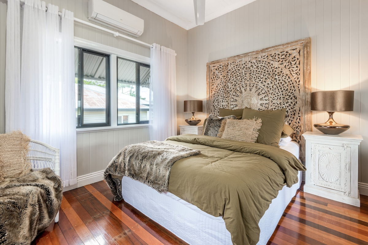 A serene bedroom is highlighted by a patterned wood feature wall above a spacious bed draped in earthy linens. Soft, natural light filters in through the large windows with sheer curtains, complementing the warm wooden floors and cozy seating area.