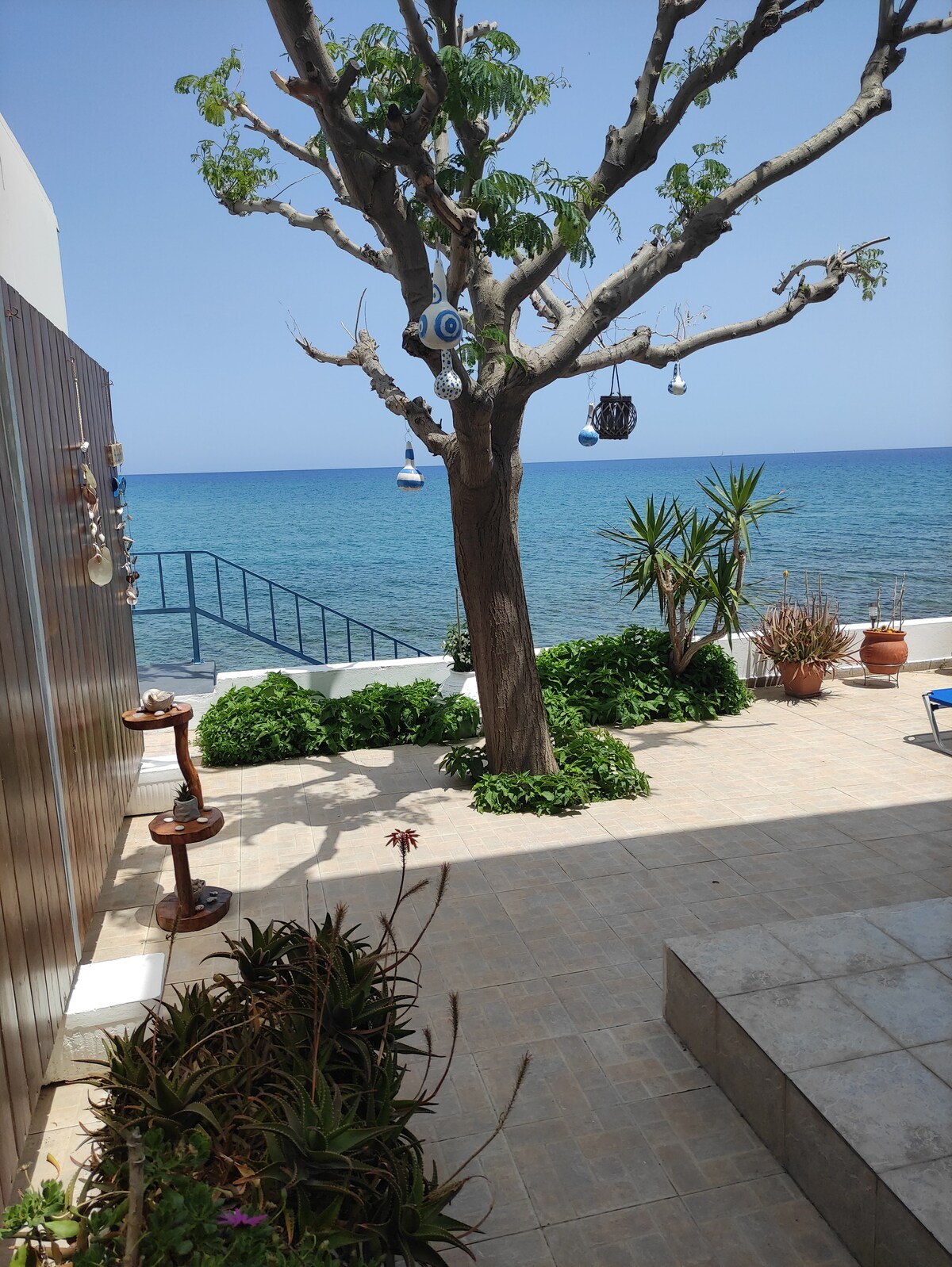 A large courtyard extends toward the sea, with a view of clear blue water visible. A tree with hanging decorations provides shade, while surrounding greenery adds a natural touch. A pathway leads down to the water, enhancing the tranquil outdoor setting.