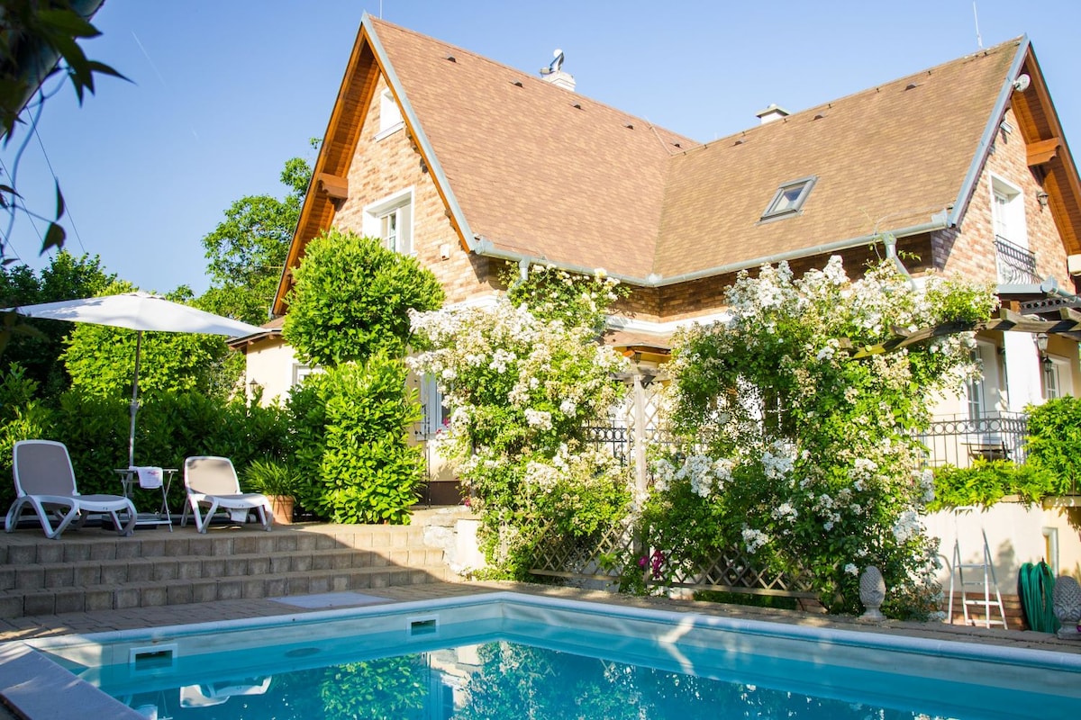 A charming house with a brown shingle roof is surrounded by lush greenery and flowering vines. A blue saltwater pool sits in the foreground, complemented by white lounge chairs and a large umbrella for shade. Sunlight illuminates the inviting scene.